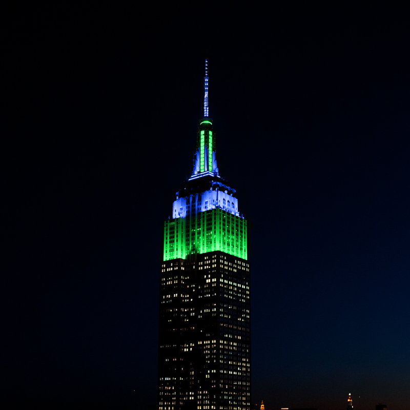 The top of the Empire State Building, lit in green and blue against the night sky.